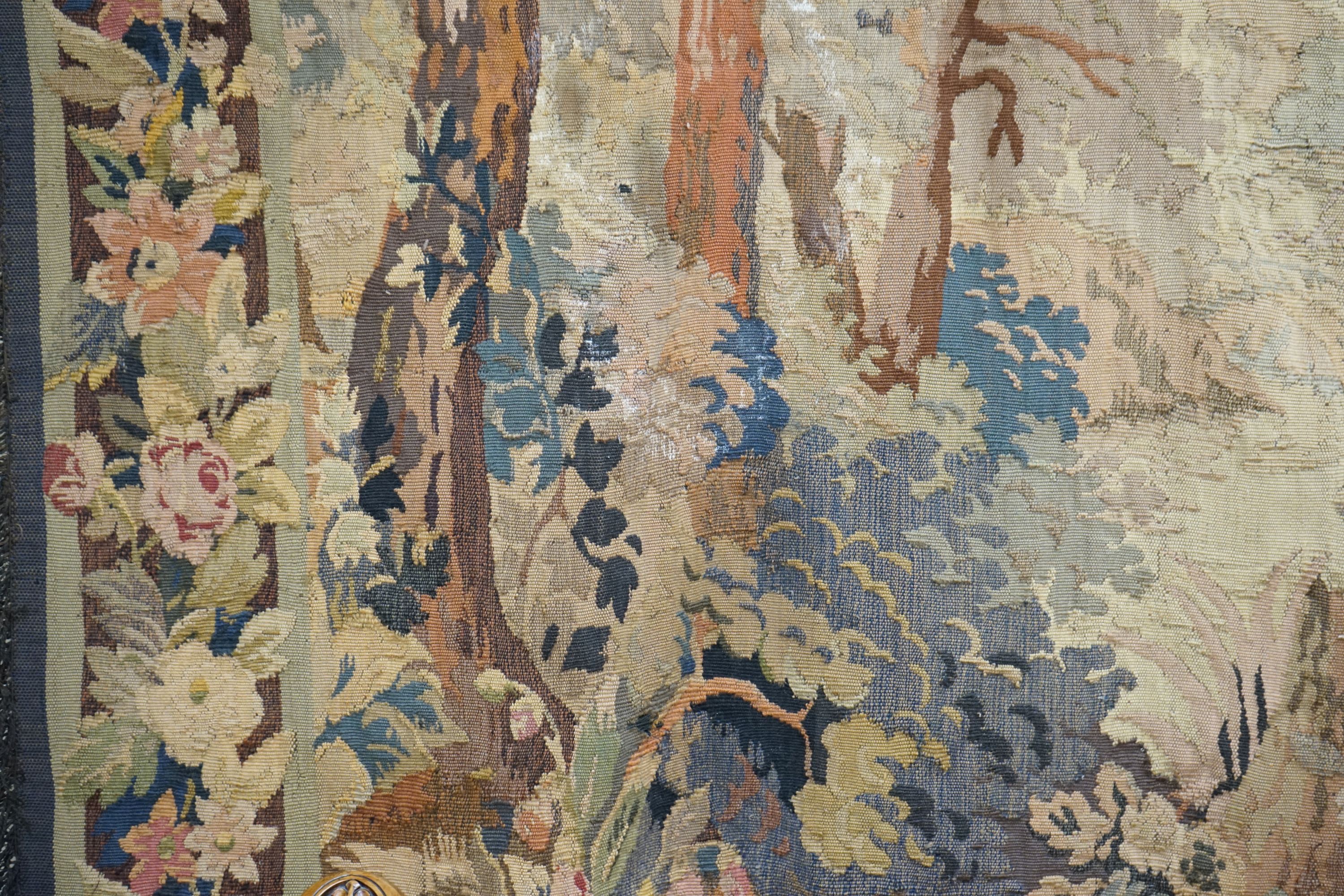 A late 19th / early 20th century French tapestry depicting a woodland scene with trees and flowers with a floral border woven in autumnal colours